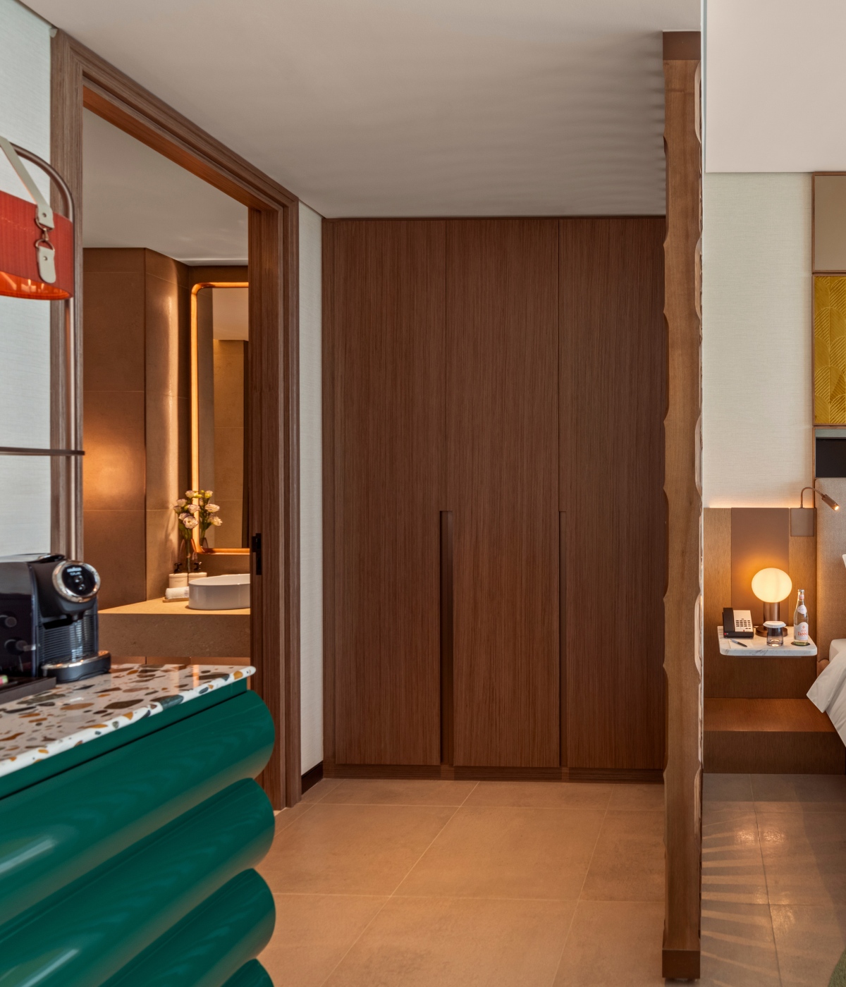 Modern hotel room corner with wooden closet, green textured counter with coffee machine, warm-lit bathroom with flowers, and cozy bedside lamp.