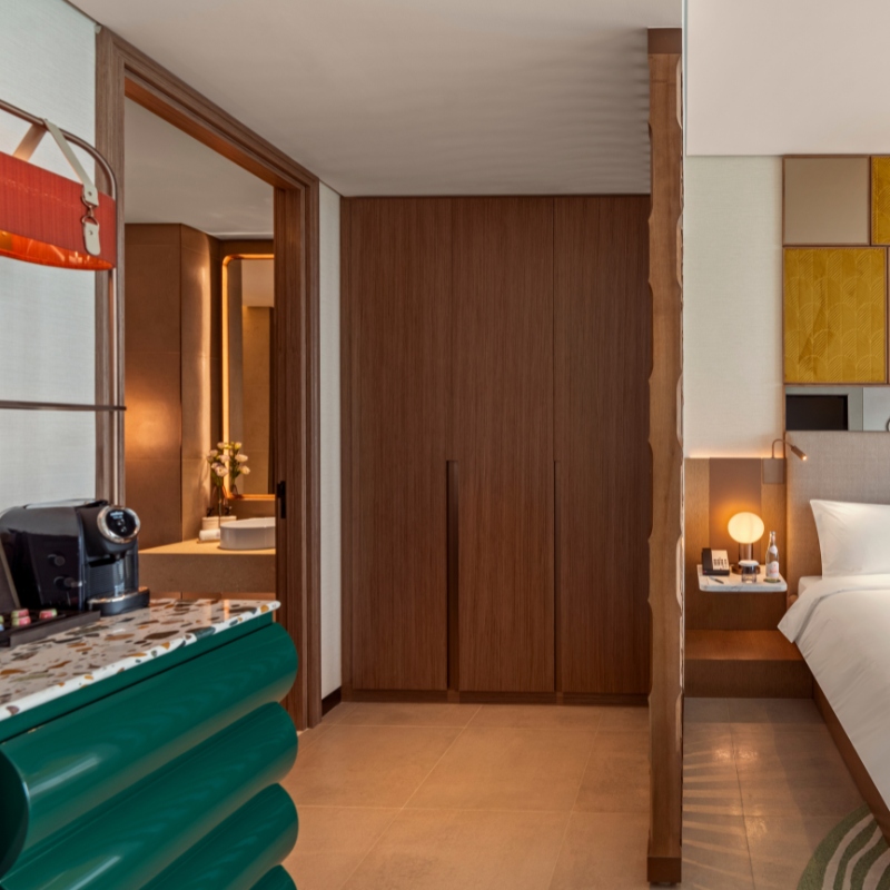 Hotel room corner with a green counter and coffee machine. Wood doors lead to a bathroom; a cozy bed with a lit lamp is partially visible, creating a warm ambiance.