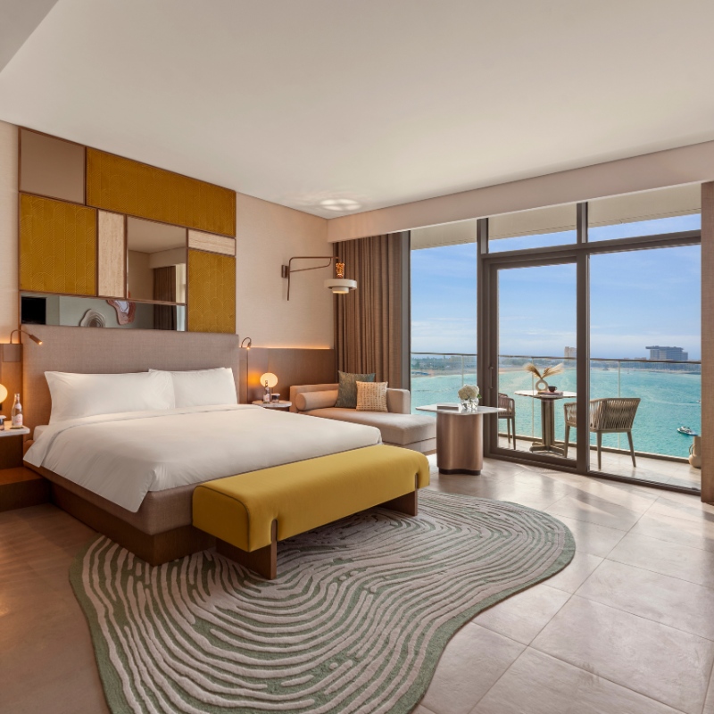 Elegant hotel room with a large bed, yellow bench, and stylish decor. Floor-to-ceiling windows reveal a stunning ocean view and a cozy balcony setup.