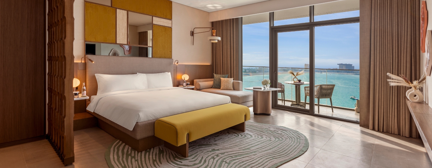 Luxurious hotel room with a large bed, yellow accents, and modern decor. Floor-to-ceiling windows offer a stunning ocean view, with a balcony seating area.