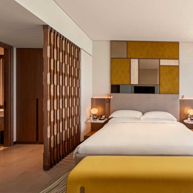 Modern hotel room with a king-sized bed, white linens, and a decorative headboard. Warm lighting and a wooden partition create a cozy, inviting atmosphere.