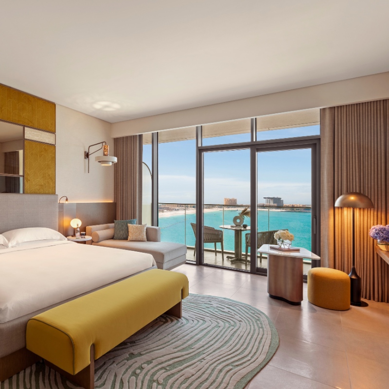 Luxurious hotel room with modern decor, featuring a spacious bed, cozy seating, and a glass door opening to a balcony. The ocean view enhances a serene, inviting ambiance.