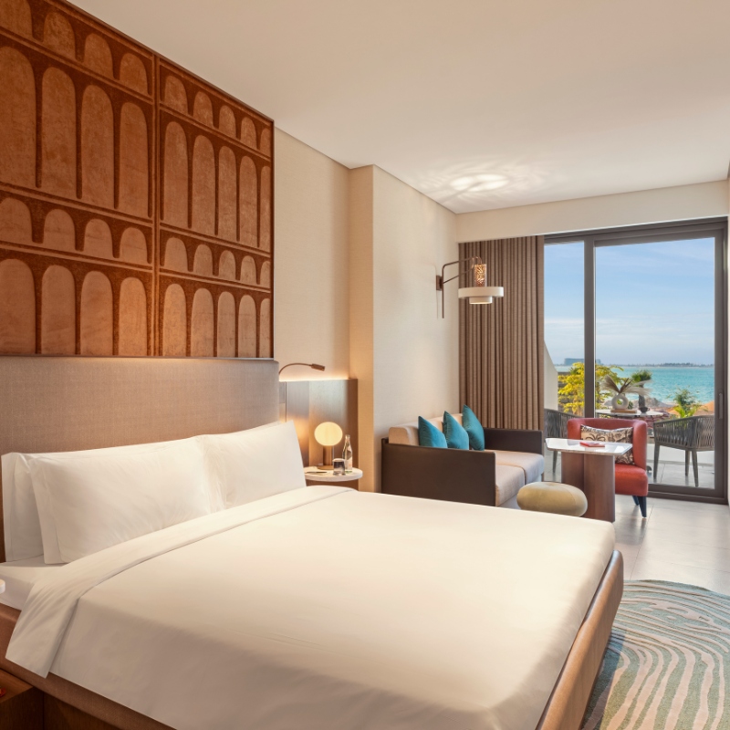 Modern hotel room with a large bed, intricate wooden wall art, and sea view. A cozy sitting area near a balcony enhances relaxation. Bright, inviting ambiance.