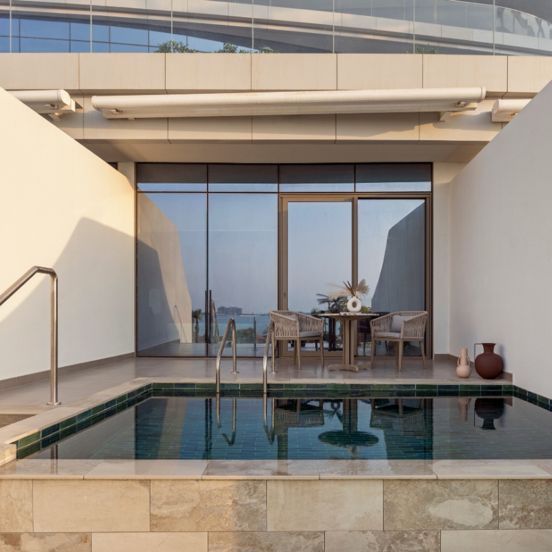 A serene patio features a small pool with a metal handrail, adjacent to glass doors leading inside. A table with chairs and a vase adds elegance. Soft lighting and ocean view create a tranquil atmosphere.