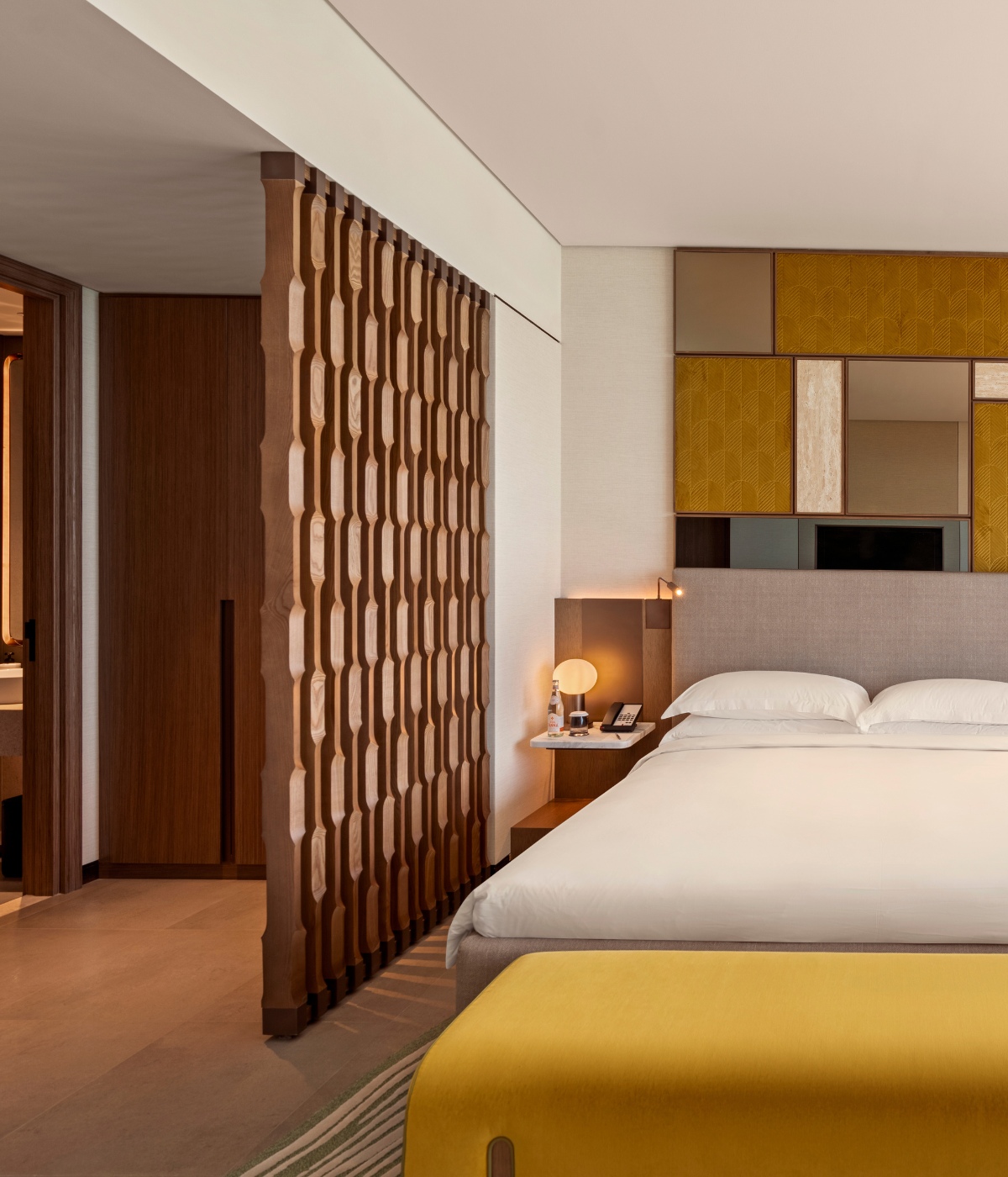 Modern hotel room with a king-sized bed, white linens, and a decorative headboard. Warm lighting and a wooden partition create a cozy, inviting atmosphere.