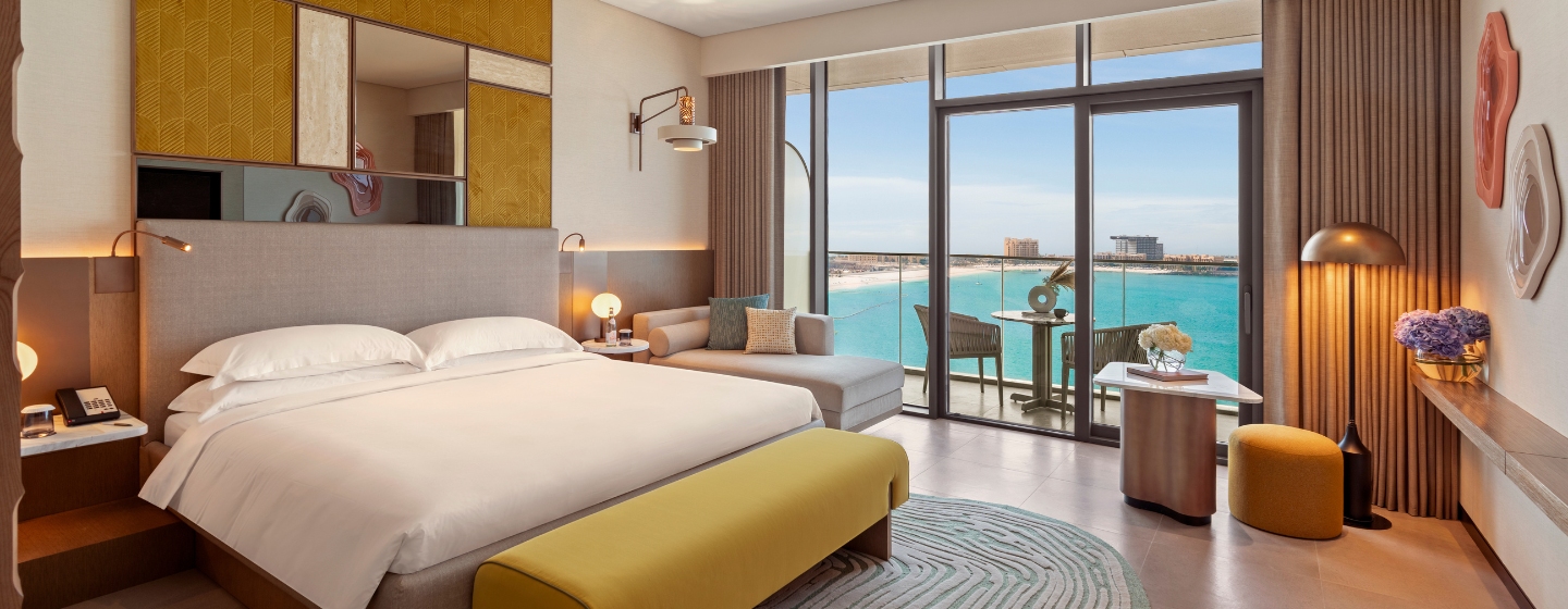 Luxurious hotel room with modern decor, featuring a spacious bed, cozy seating, and a glass door opening to a balcony. The ocean view enhances a serene, inviting ambiance.