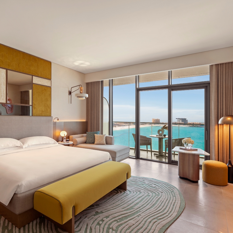 Luxurious hotel room with modern decor, featuring a spacious bed, cozy seating, and a glass door opening to a balcony. The ocean view enhances a serene, inviting ambiance.