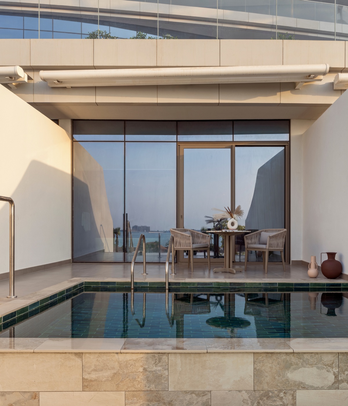 A serene patio features a small pool with a metal handrail, adjacent to glass doors leading inside. A table with chairs and a vase adds elegance. Soft lighting and ocean view create a tranquil atmosphere.
