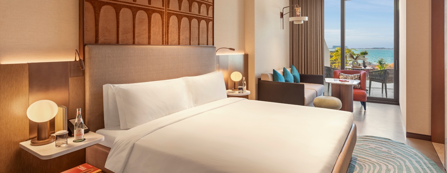 Modern hotel room with a large bed, soft lighting, and a view of the ocean through a glass door. Cozy seating area with a sofa and colorful pillows.