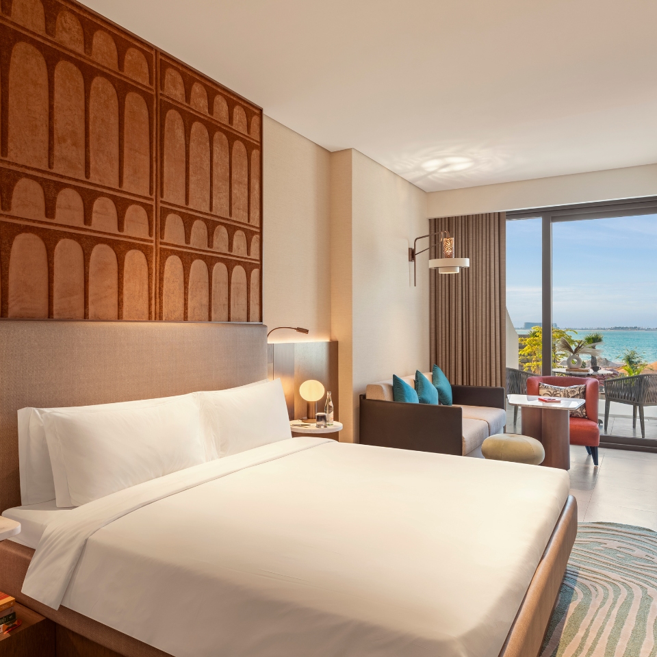 Modern hotel room with a large bed, soft lighting, and a view of the ocean through a glass door. Cozy seating area with a sofa and colorful pillows.