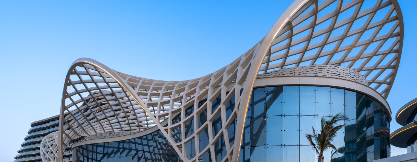 Futuristic building with a wavy, lattice-like structure on its roof, set against a clear blue sky. Reflective glass facade, with palm trees nearby.