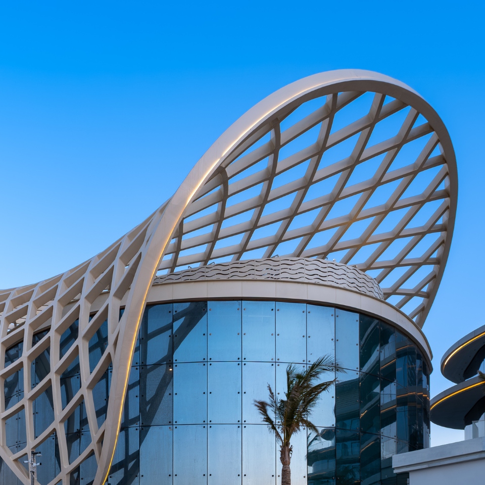 Futuristic building with a wavy, lattice-like structure on its roof, set against a clear blue sky. Reflective glass facade, with palm trees nearby.
