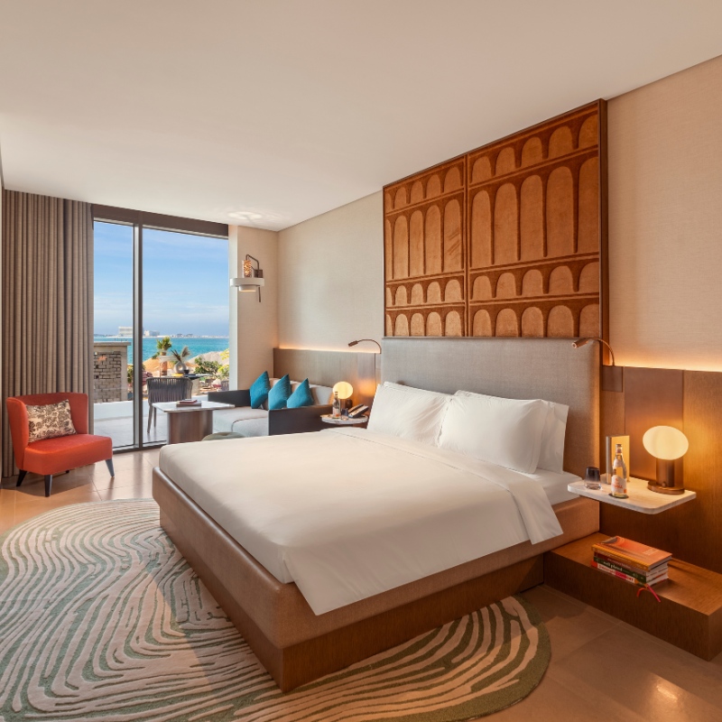 Stylish hotel room with ocean view. Features a large bed, modern wood paneling, art deco rug, soft lighting, and a window with a bright, scenic beach view.