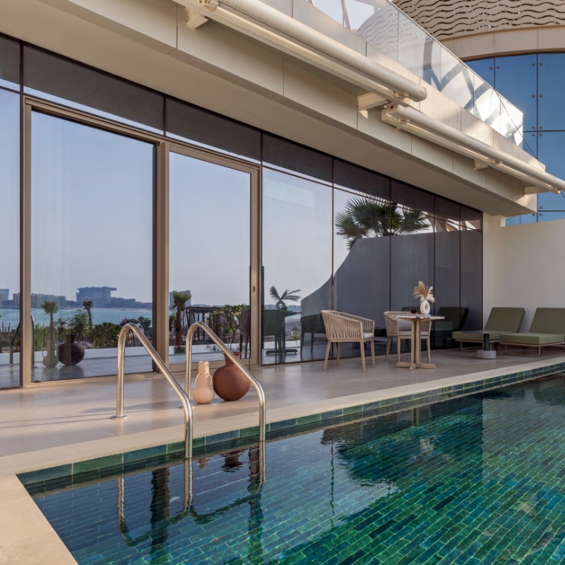 Luxurious outdoor poolside with clear water reflecting light. Modern lounge area with wicker chairs, table, and a scenic city view in the background.