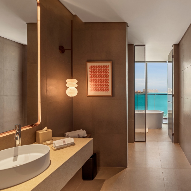 Modern bathroom with neutral tones featuring a round sink on a beige counter, hanging globe light, abstract wall art, and ocean view with freestanding tub.