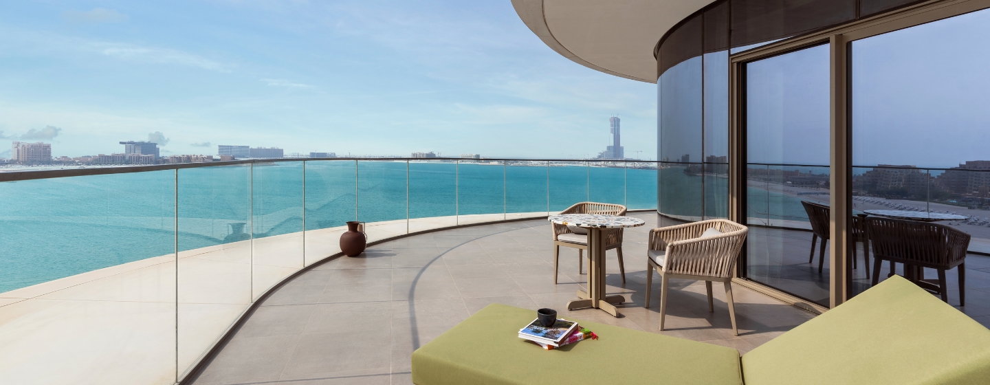 Curved balcony overlooking a calm blue ocean, featuring a green lounge chair and table with chairs. A serene, modern setting under a clear sky.