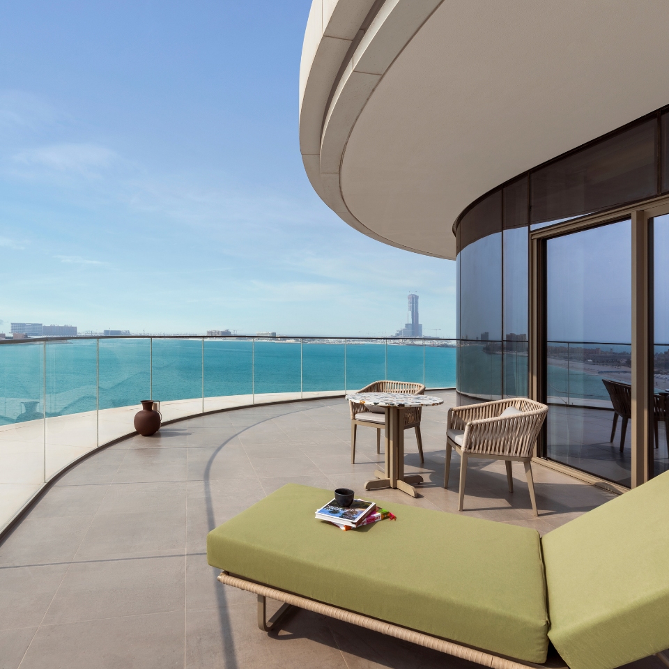 Curved balcony overlooking a calm blue ocean, featuring a green lounge chair and table with chairs. A serene, modern setting under a clear sky.