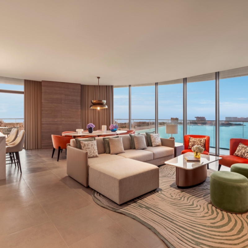 Modern living room with a large beige sectional sofa and a sleek white coffee table on a patterned rug. Floor-to-ceiling windows offer an ocean view. A dining area with orange chairs and decorative flowers adds warmth. The atmosphere is serene and elegant.