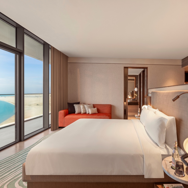 A modern hotel room with a large bed, white linens, and elegant wooden decor. A floor-to-ceiling window offers a view of a serene beach and ocean.