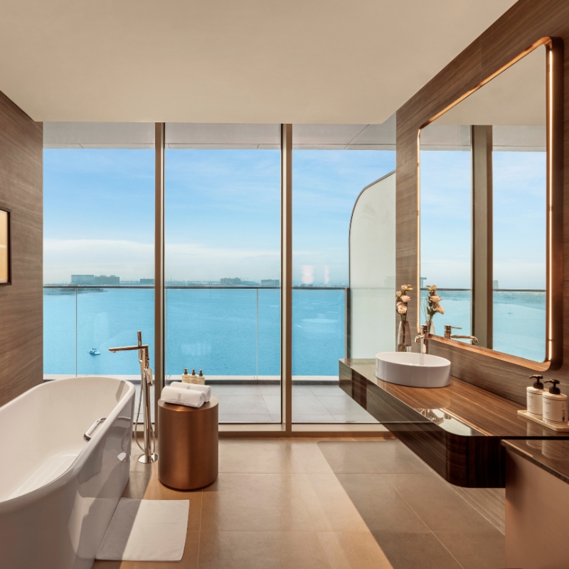 Luxurious bathroom with a freestanding tub, large mirror, and vessel sink. Floor-to-ceiling windows offer a stunning ocean view under a clear sky.