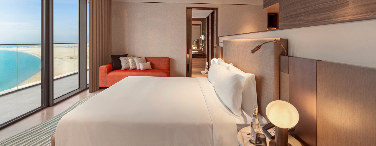 A modern hotel room with a large bed, white linens, and elegant wooden decor. A floor-to-ceiling window offers a view of a serene beach and ocean.