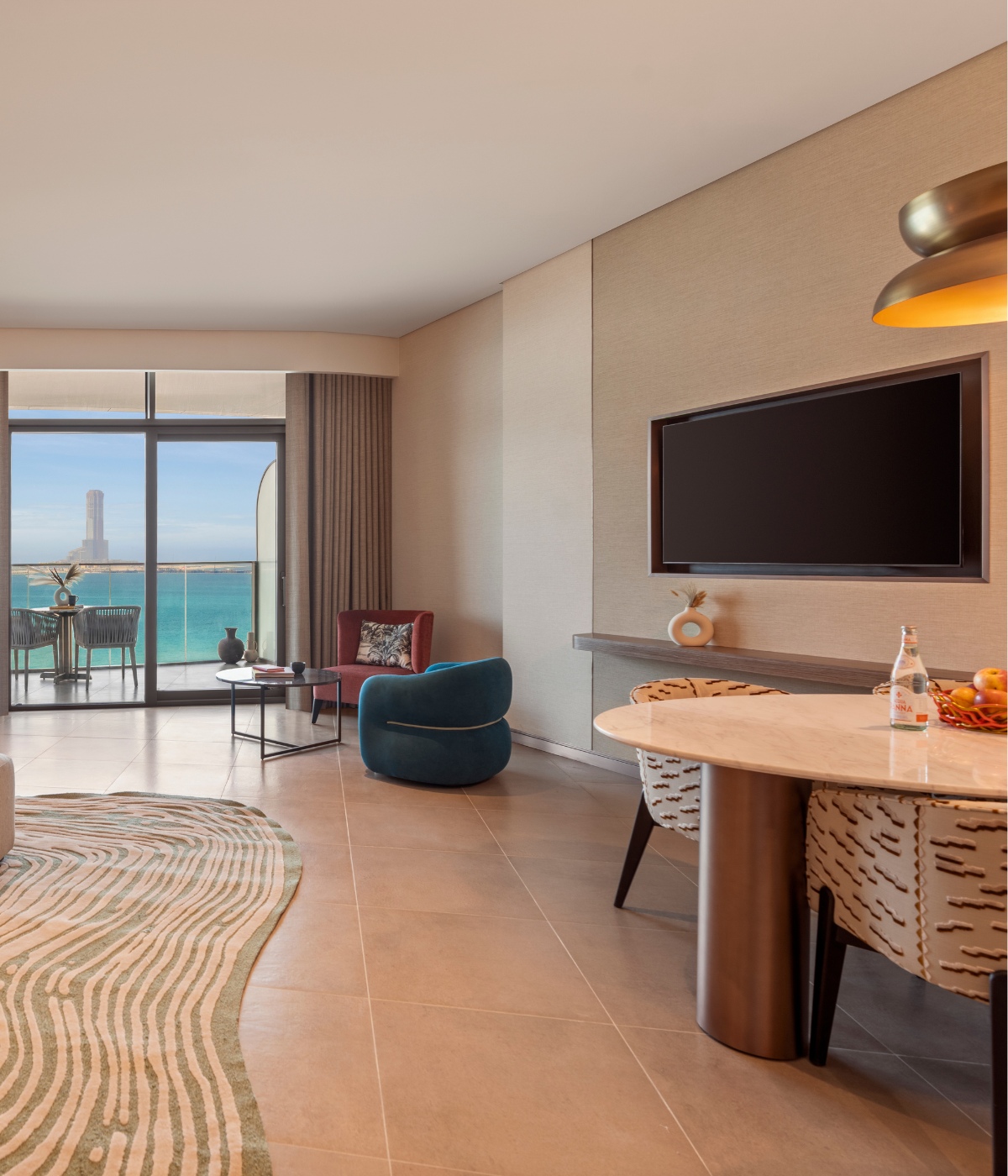 Modern living room with a sea-view balcony. Features a teal chair, lamp-lit round table with fruit, TV, and wavy-patterned rug. Warm and inviting ambiance.