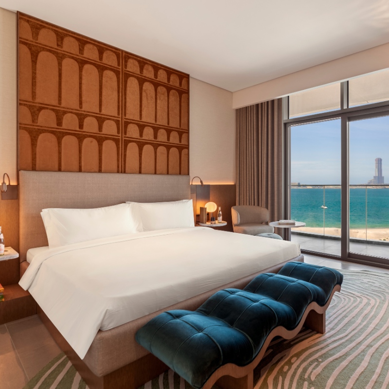 Modern hotel room with a large bed featuring a patterned headboard, turquoise benches, and a view of the ocean through floor-to-ceiling windows.