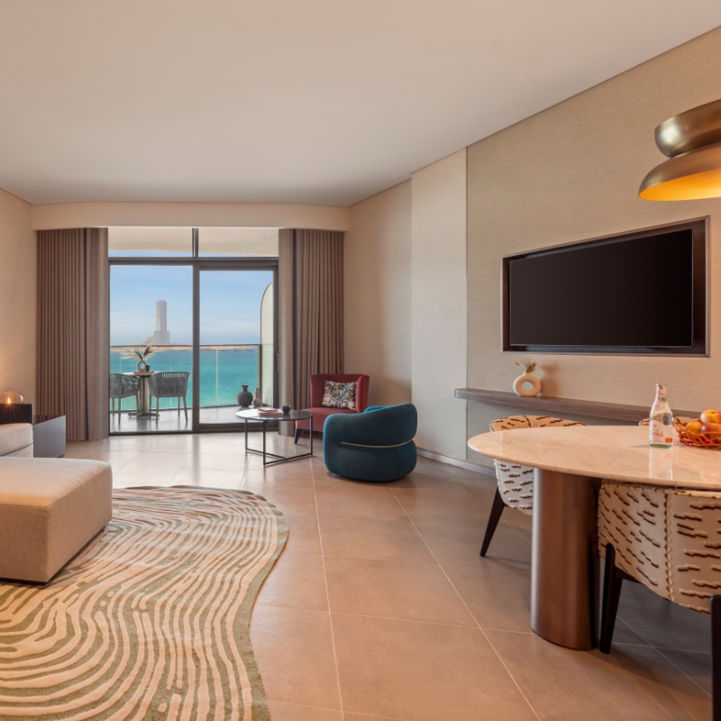 Modern living room with a sea-view balcony. Features a teal chair, lamp-lit round table with fruit, TV, and wavy-patterned rug. Warm and inviting ambiance.