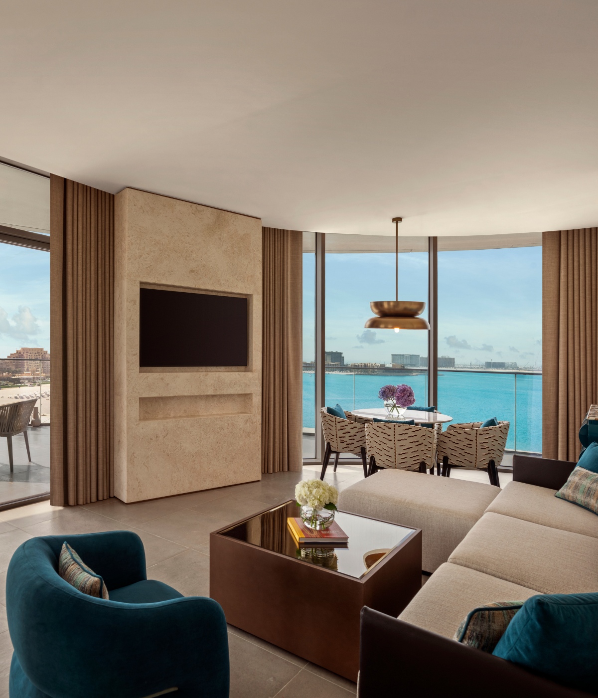 Modern living room with large windows overlooking a blue ocean. Features a beige sofa, blue-green chairs, wall-mounted TV, and a dining table with flowers.