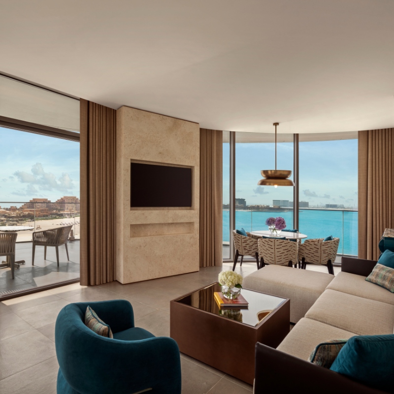 Modern living room with large windows overlooking a blue ocean. Features a beige sofa, blue-green chairs, wall-mounted TV, and a dining table with flowers.