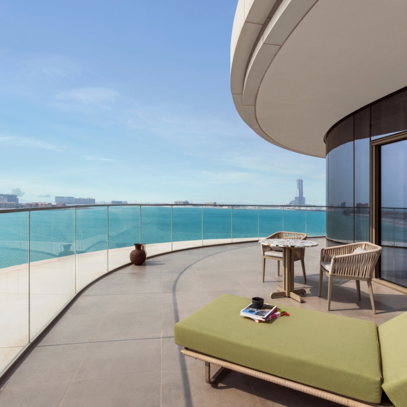 Spacious balcony overlooking a calm, blue ocean with clear skies. Features a green cushioned lounger, a round table with chairs, and a serene, relaxing ambiance.