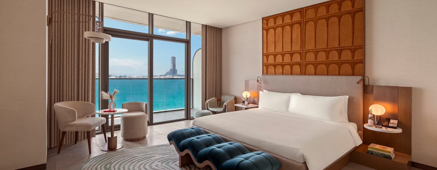 Modern hotel room with a king-sized bed, elegant decor, and ocean view through large windows. Calm tones, plush seating, and cozy atmosphere.