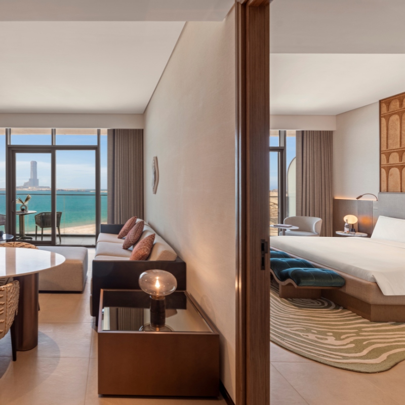 Modern hotel suite with ocean view; living area with sofa, cushions, lamp, and dining table. Bedroom features large bed, artistic rug, warm ambiance.