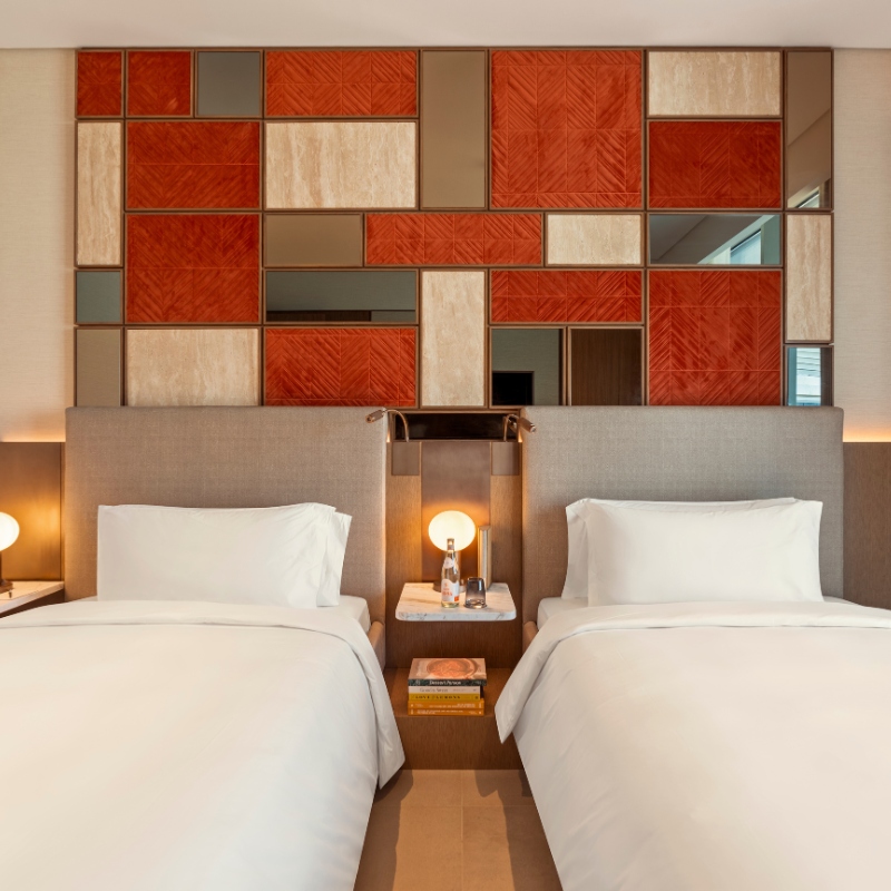 Modern hotel room with two neatly made beds, warm wooden nightstand between them. Decorative red and beige geometric wall panel adds elegance.
