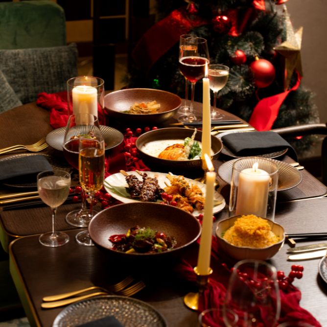 Elegant holiday table setting with candlelit ambiance. Plates of gourmet dishes and glasses of wine are arranged on a dark wood table.