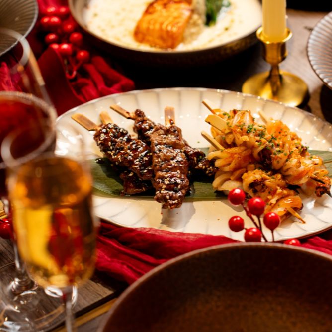 Festive table setting with skewers of glazed meat and seasoned fried potatoes on a ceramic plate. Red berries, a candle, and champagne add elegance.