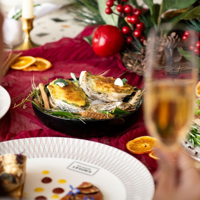 Festive table setting with baked oysters in a black dish, garnished with greens. A glass of champagne is in the foreground, and the table is decorated with red berries, pine cones, and orange slices.