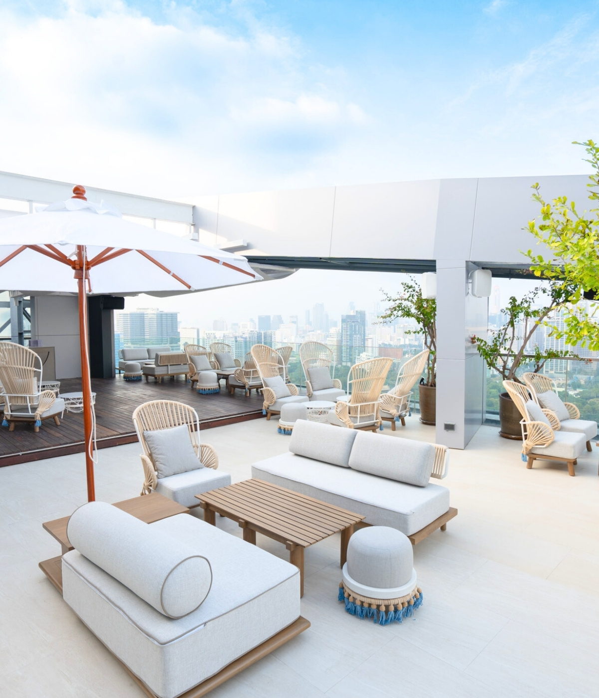Rooftop terrace with chic white and beige seating, a large umbrella, and lush plants. Offers city skyline views under a clear, sunny sky.