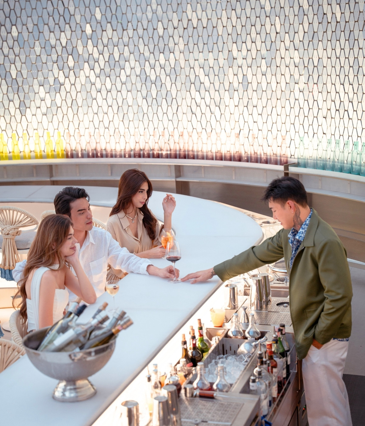 A stylish bar setting with three people seated, two women and a man, engaging with the bartender. Glasses of wine are on the counter, under soft lighting.