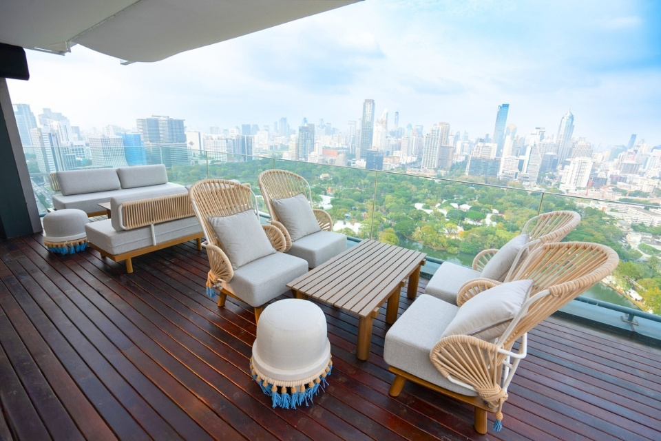 http://Luxurious%20rooftop%20terrace%20with%20wicker%20furniture%20and%20soft%20cushions%20on%20wooden%20decking,%20overlooking%20a%20vibrant%20cityscape%20and%20lush%20park%20under%20a%20blue%20sky.