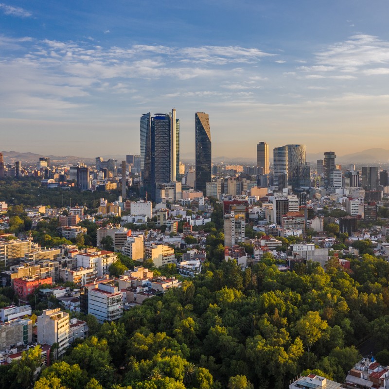 Aerial view of a bustling cityscape with tall skyscrapers and modern buildings under a blue sky. Green park areas are in the foreground, adding contrast.