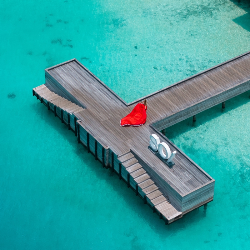 Aerial view of a wooden pier shaped like a cross over clear turquoise water. A person in a flowing red dress lies on the pier, creating a vivid contrast.