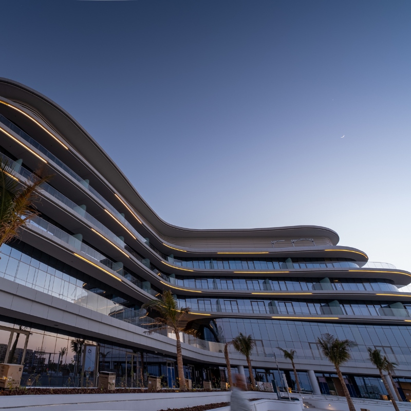 Modern multi-story building with curved architecture, large glass windows, and palm trees, illuminated under a twilight sky.