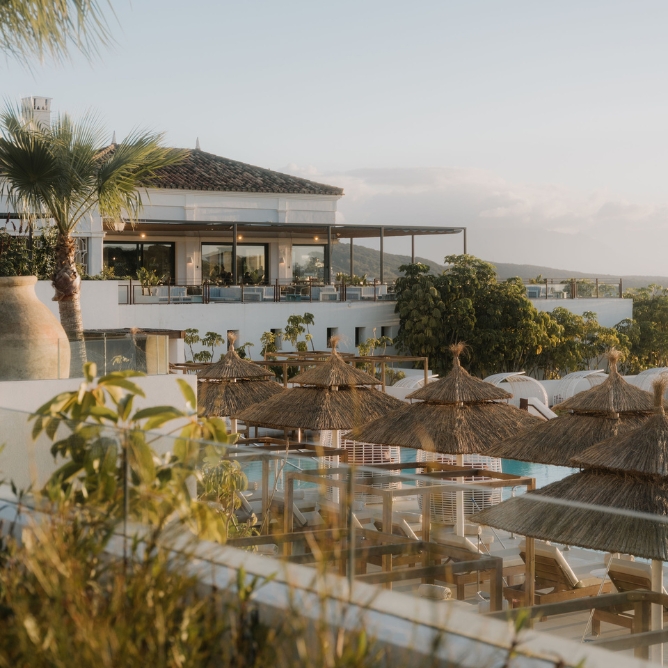 Scenic view of a modern resort with a rooftop terrace, lush greenery, and thatched umbrellas beside a tranquil pool under a clear sky.