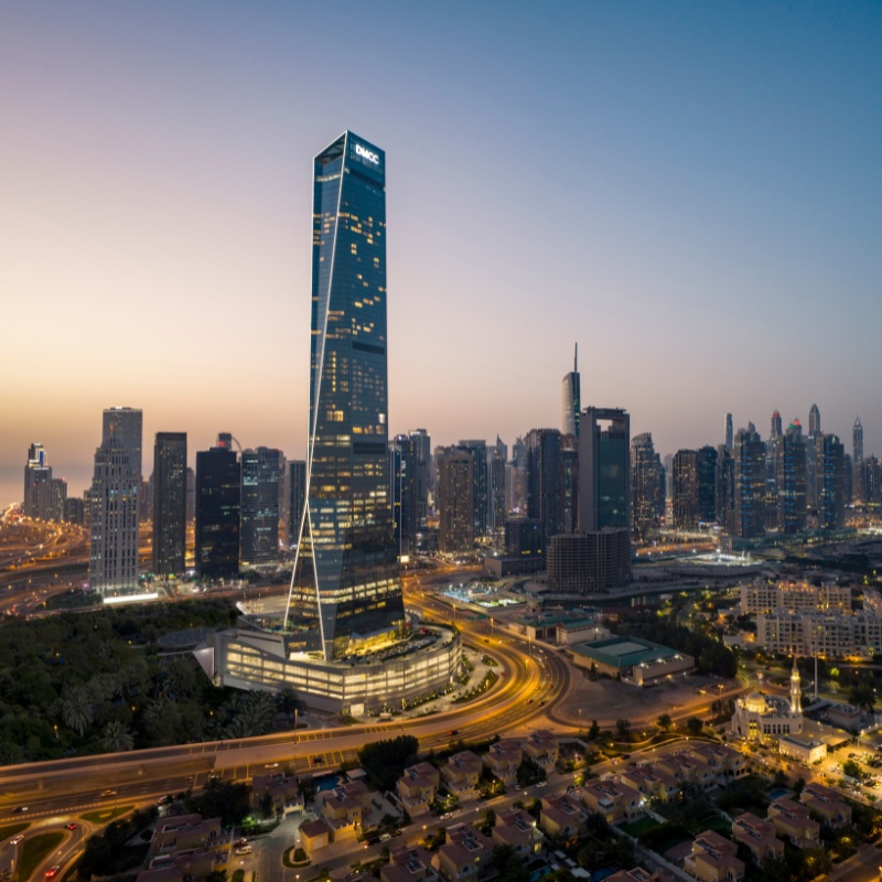Aerial view of Dubai at dusk, showcasing a tall, illuminated skyscraper amidst a glowing urban skyline. Roads curve below, hinting at a bustling city.
