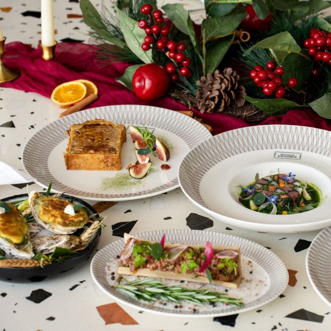 Elegant holiday table setting with gourmet dishes, including oysters, beef tartare, figs, and greens. Festive decor with red berries and candles.