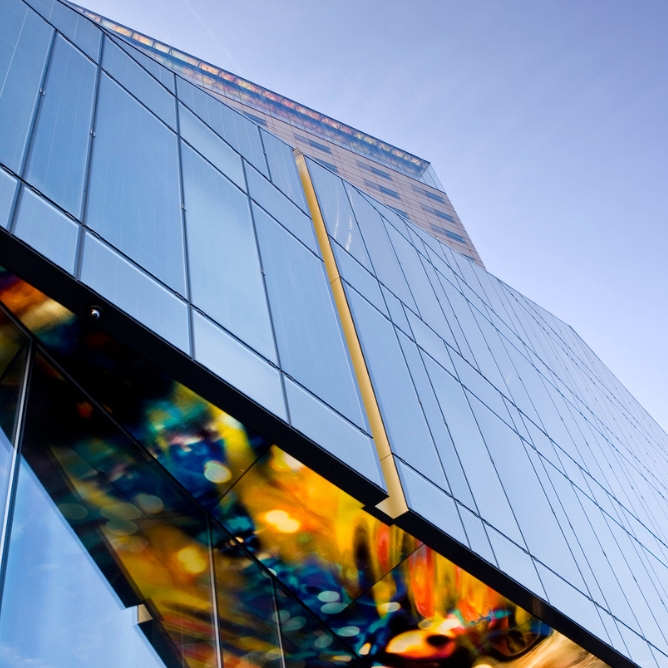 Looking up at a modern glass building adorned with colorful artwork on its base, against a clear blue sky.