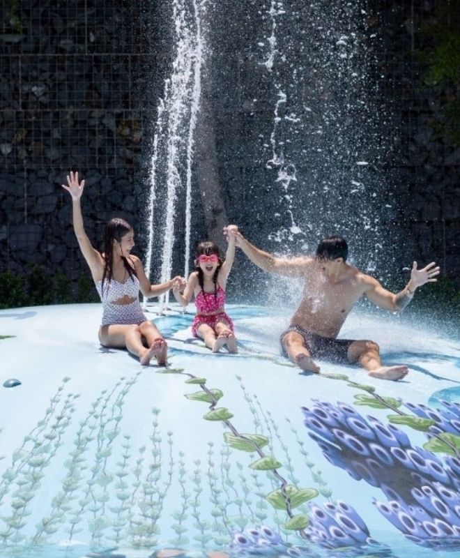 Three people joyfully playing in a water park; one on a yellow float, two others standing and jumping amidst splashing water, all smiling brightly.