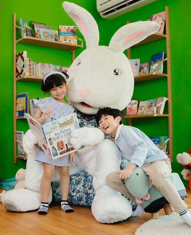 A young girl reading a book and a boy laughing next to a giant white rabbit stuffed animal in front of green bookshelves at the Kids Tent, SO/ Sofitel Hua Hin