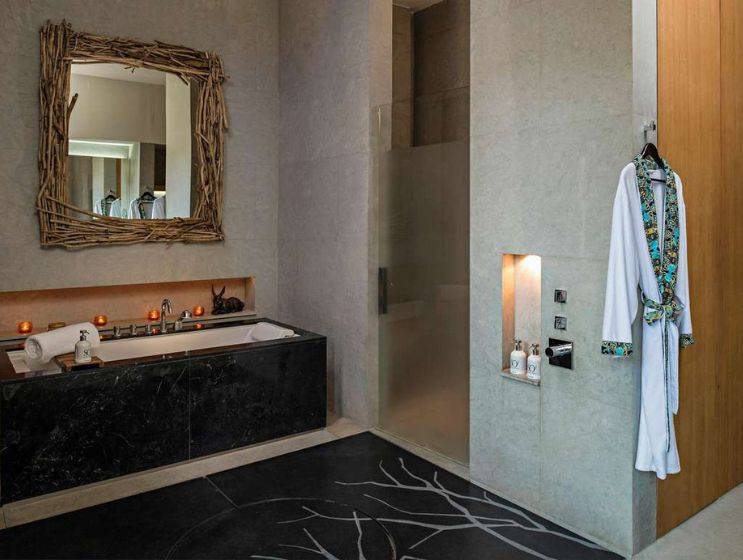 A modern spa bathroom with a deep soaking tub surrounded by dark marble, a walk-in shower with a glass panel, and a unique mirror framed with natural wood branches.